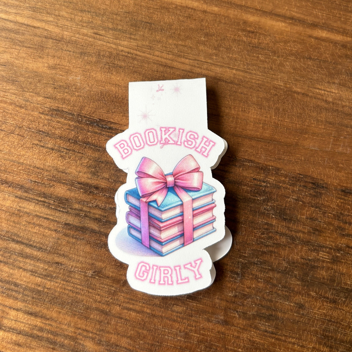 magnetic bookmark with 'Bookish Girly' text and a stack of books with a pink bow on a wooden surface.