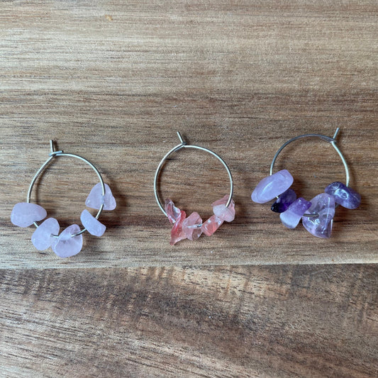 A collection of silver wine glass charm rings with various gemstones attached to them, displayed on a wooden surface.