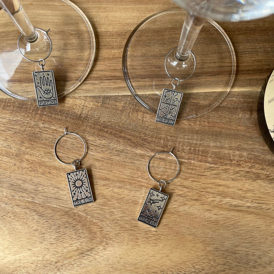 Four silver wine glass charms with tarot card charms attached, displayed on a wooden surface next to a glass of wine.