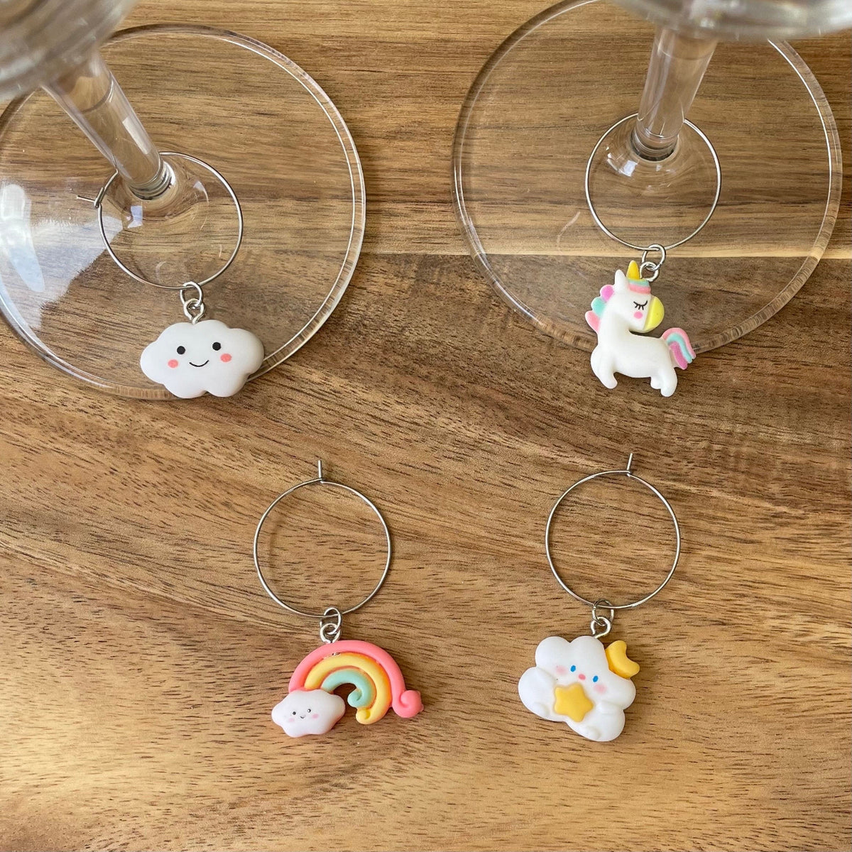 A set of four silver wine glass charm rings with various resin charms including a rainbow, unicorn, cloud, and cloud with star and moon, displayed on a wooden surface alongside two wine glasses.