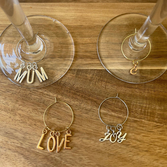 A collection of silver and gold wine glass charm rings with gold and silver capital letters and cursive letters attached to them, displayed on a wooden surface