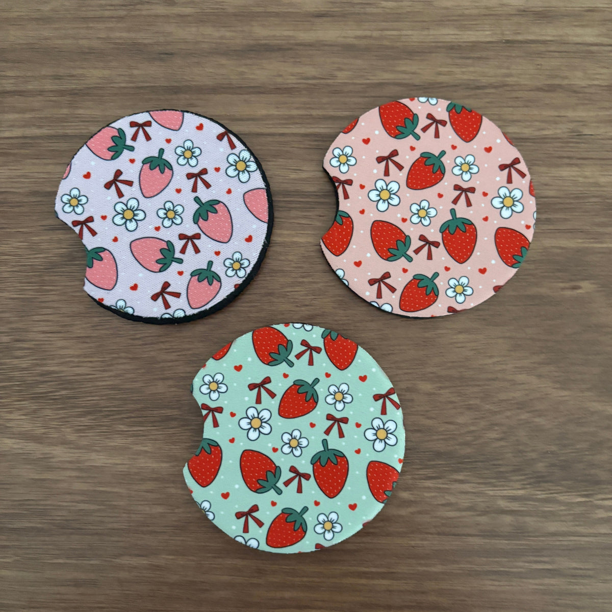 Three circular fabric coasters with curved cutouts, featuring strawberry and flower patterns in pink, light purple, and mint green, arranged on a brown wooden surface.