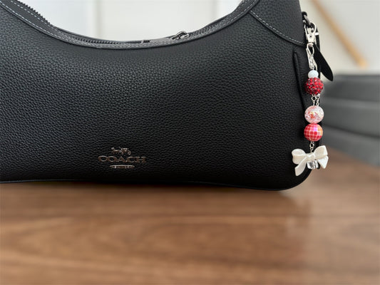 A close-up of a black pebbled leather Coach shoulder bag resting on a wooden surface, featuring a decorative charm with red and white patterned beads and a white bow.