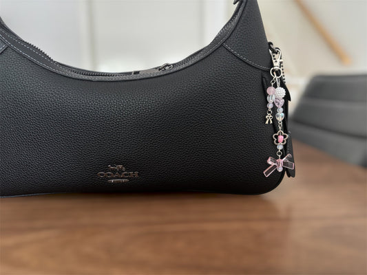 A close-up of a black pebbled leather shoulder bag with a silver-tone metal logo plate on the front and a dangling charm strap with pink and silver decorative elements, resting on a wooden surface.