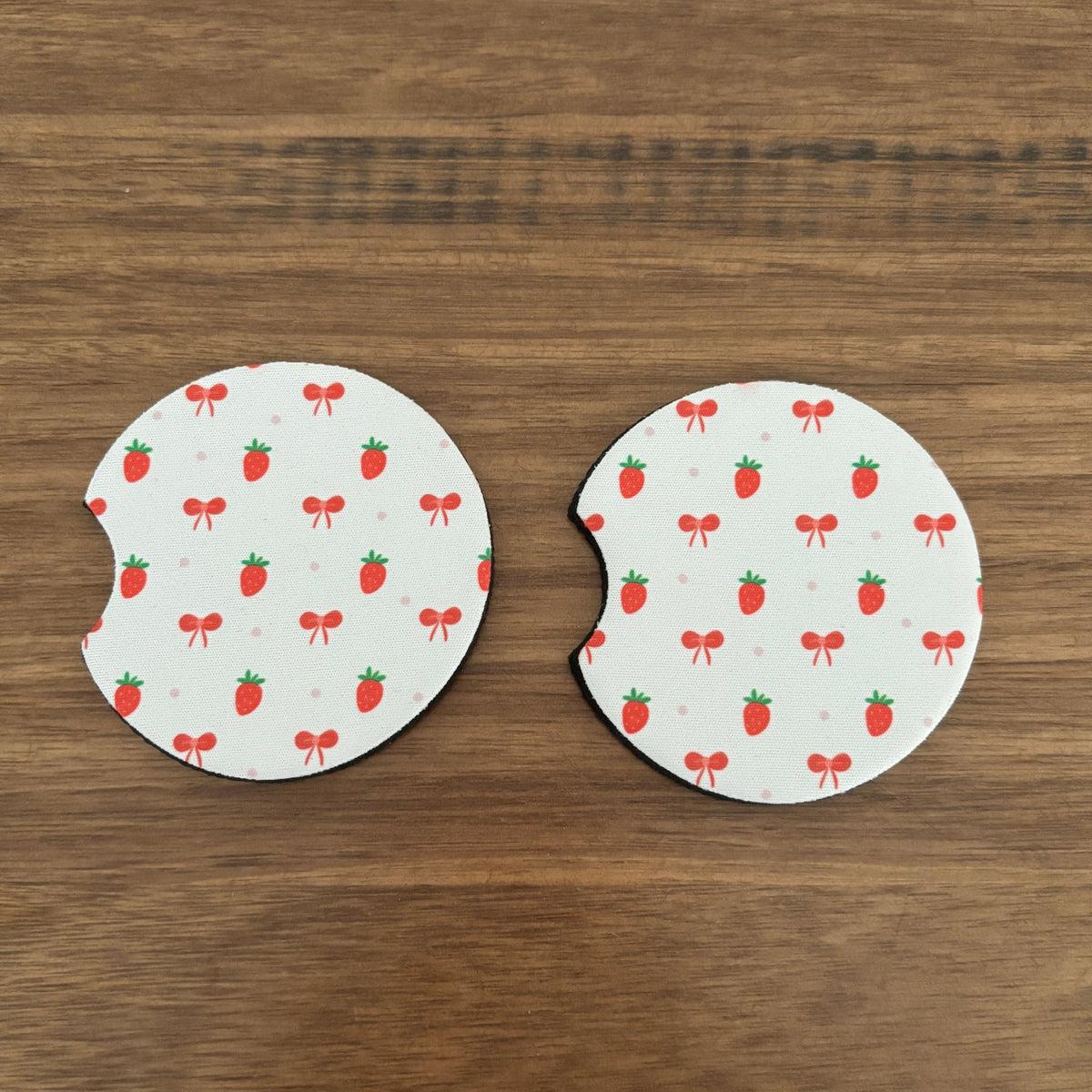 Two circular white coasters with a strawberry and bow pattern, shown on a brown wooden surface.