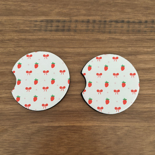Two circular white coasters with a strawberry and bow pattern, shown on a brown wooden surface.