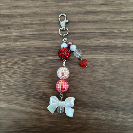 A handmade beaded charm keychain with a silver lobster clasp, featuring red rhinestone, patterned red and pink beads, a clear bead with glitter, a small red heart, and an iridescent white bow, displayed on a brown wood background.