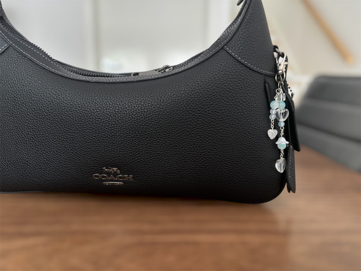 A close-up of a black pebbled leather Coach shoulder bag resting on a wooden surface, featuring a silver-tone zipper, a black leather logo patch, and a dangling charm with light blue beads and silver heart charms.