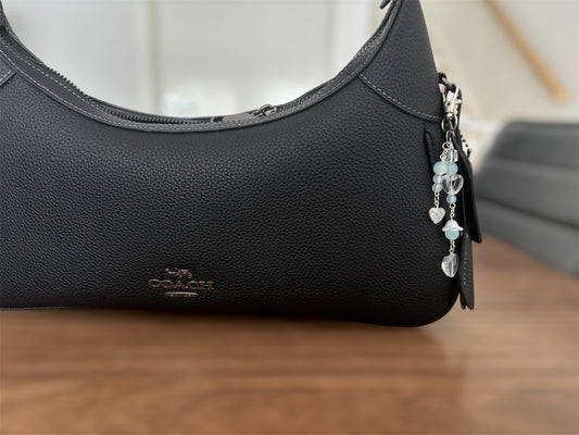 A close-up of a black pebbled leather Coach shoulder bag resting on a wooden surface, featuring a silver-tone zipper, a black leather logo patch, and a dangling charm with light blue beads and silver heart charms.