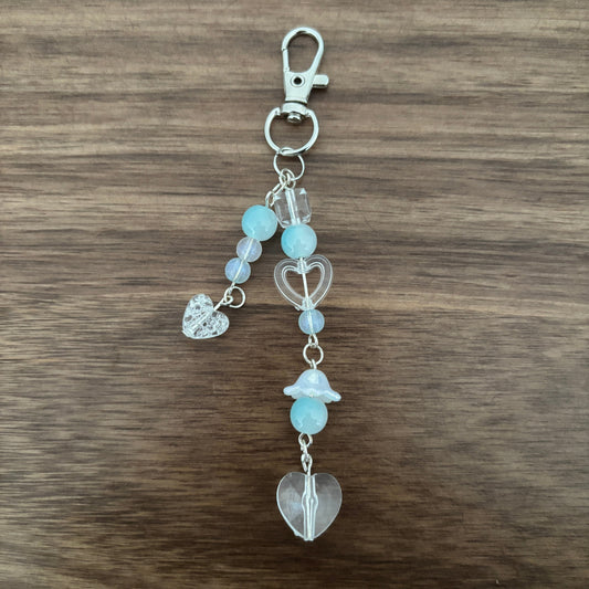 A beaded keychain charm with a silver lobster clasp, featuring light blue and lavender beads, clear heart-shaped beads, a small silver heart charm, and a larger clear heart pendant, displayed on a dark wood background.