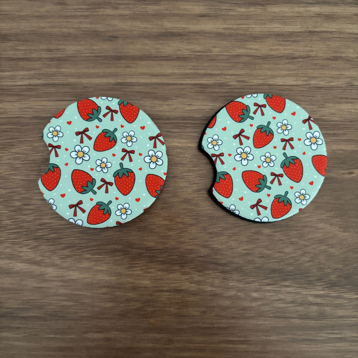 Two round coasters with a strawberry and flower pattern on a mint green background, placed on a brown wooden surface.